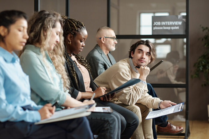 Young Man Sitting With Resume In Waiting Room Among Other Candid