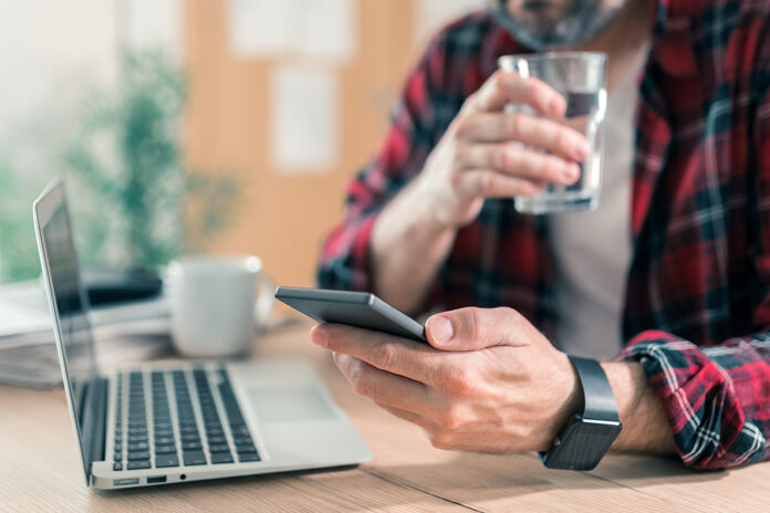 Gig Worker Drinking Water And Using Mobile Phone In Home Office