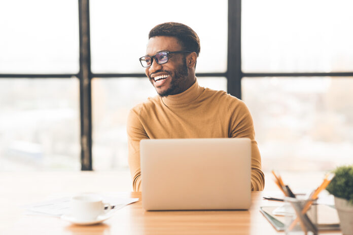 Success At Work. Happy Afro Businessman Sitting At Desk By Large
